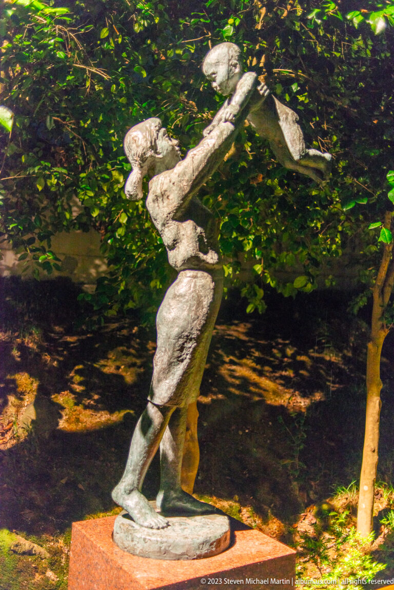 Bronze adult figure lifting a smaller figure overhead in a garden at night.