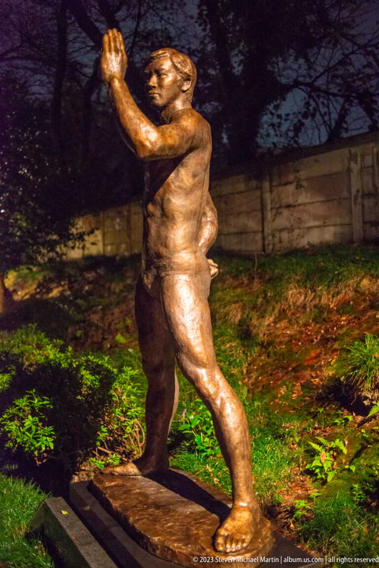 Bronze male figure twisting with one arm raised, illuminated in a garden at night.