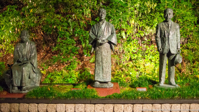 Three bronze figures in traditional and modern Japanese dress standing before a leafy wall.