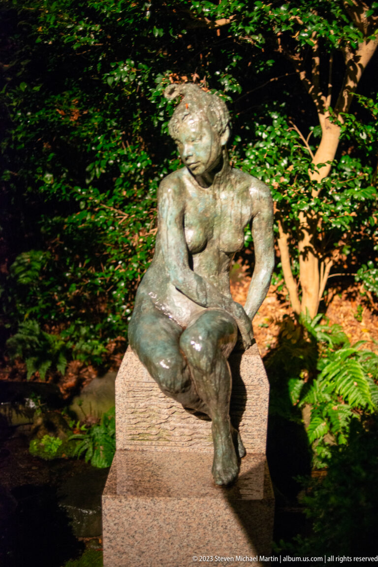 Bronze woman seated forward on a rectangular stone pedestal in a dark garden.
