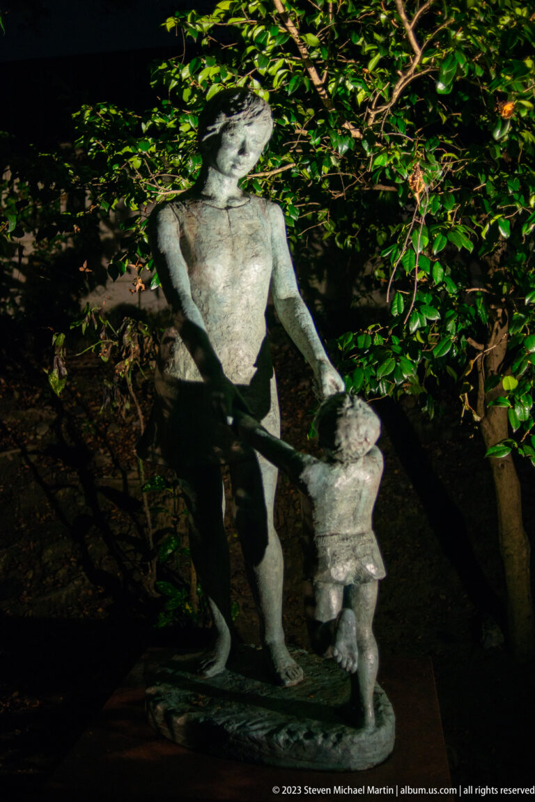 Bronze adult bending toward a small child figure beneath dense garden foliage.