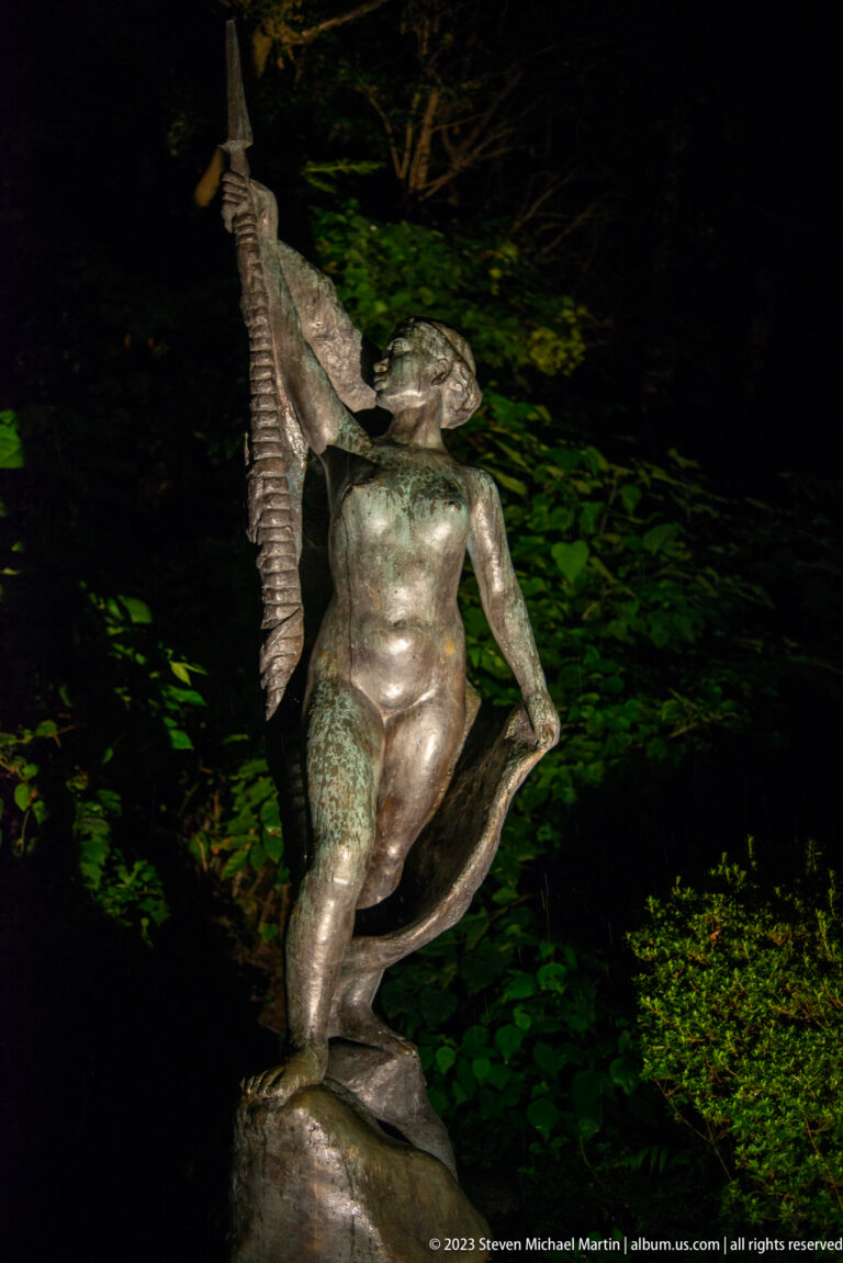 Bronze woman raising a large fish beside her shoulder in a nighttime garden.