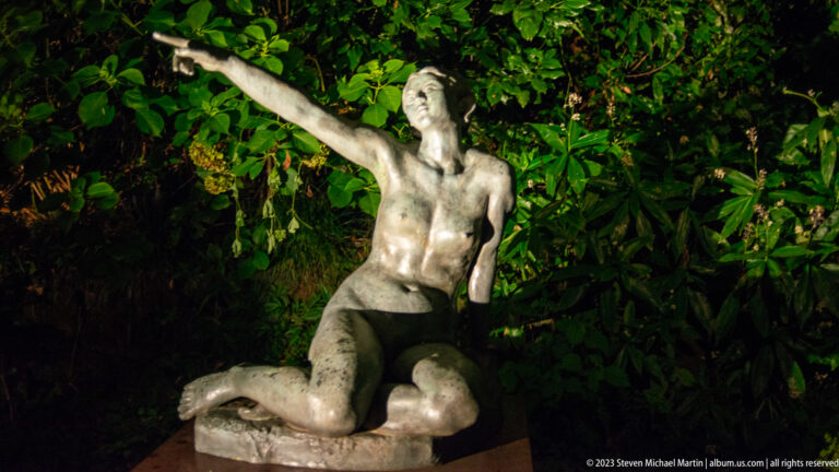 Bronze female figure reclining or kneeling with one arm extended upward against dark foliage.