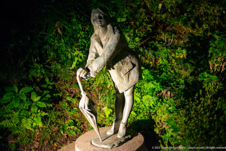 Bronze pedestrian leaning forward with a closed umbrella and shoulder bag in a garden.