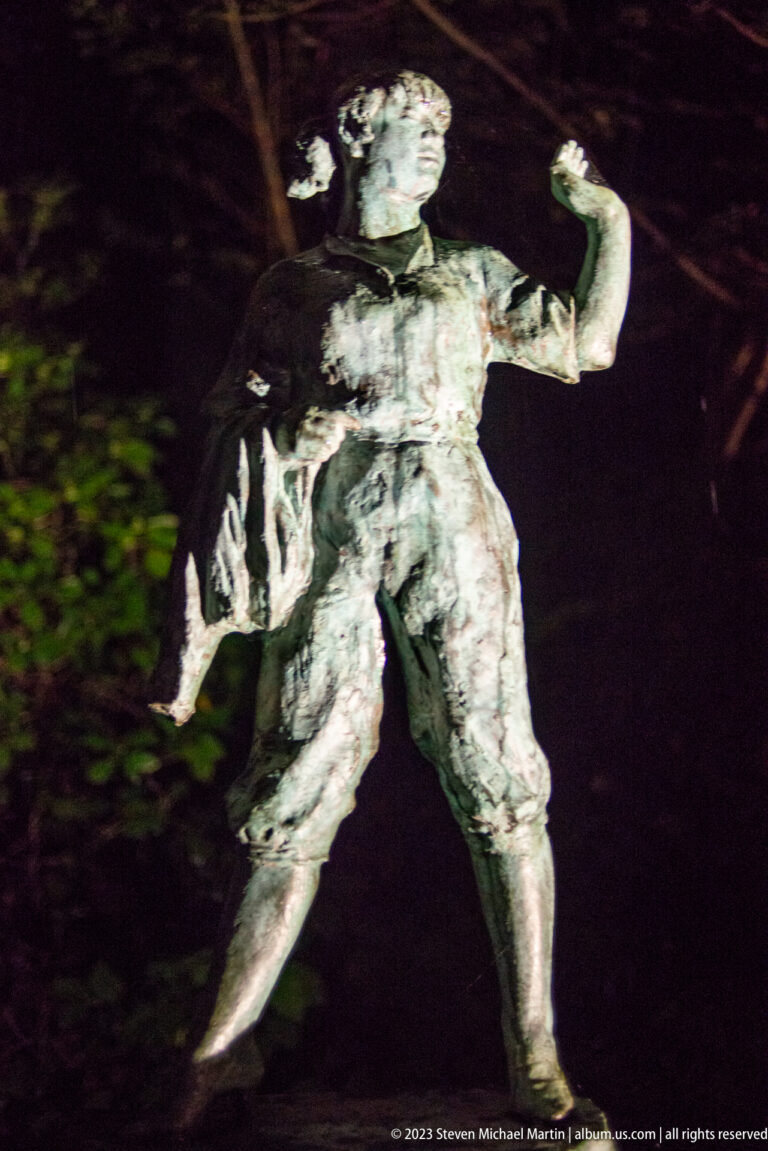 Textured male sculpture with raised arms and a wide stance, illuminated against a dark garden.