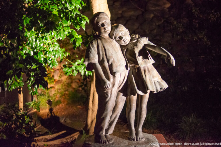 Two child figures leaning together in an illuminated outdoor sculpture surrounded by foliage.