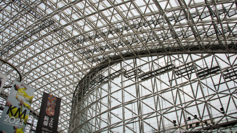 Curving steel-and-glass lattice of the Motenashi Dome with event banners hanging below.