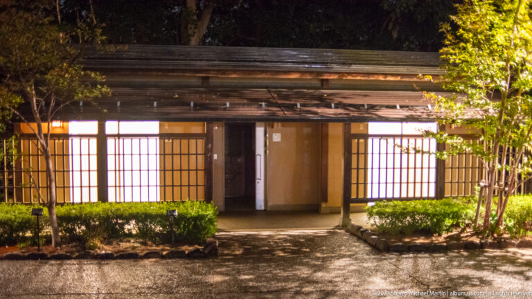 Low traditional wooden building with glowing screens, deep eaves, shrubs, and a stone path at night.