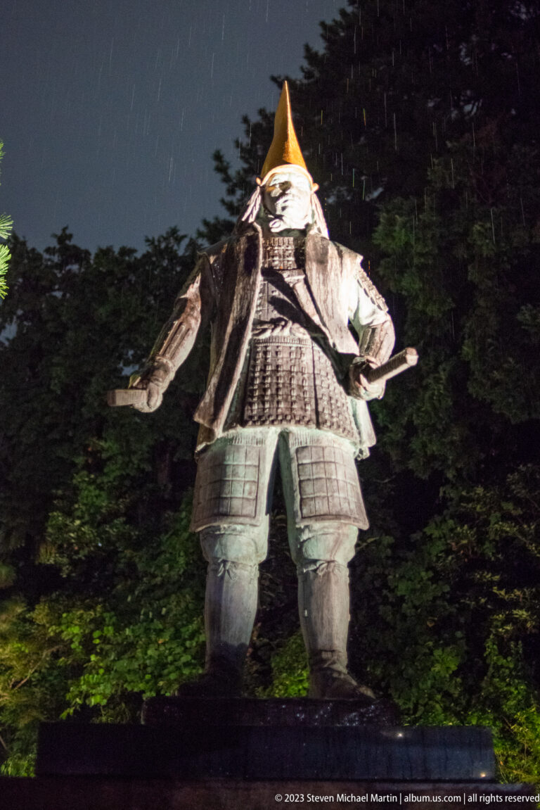 Large samurai sculpture in layered armor and a pointed helmet, illuminated from below at night.