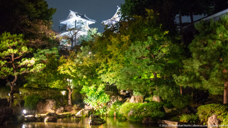 Illuminated pond garden with pines and rocks, with a white castle structure rising behind the trees.