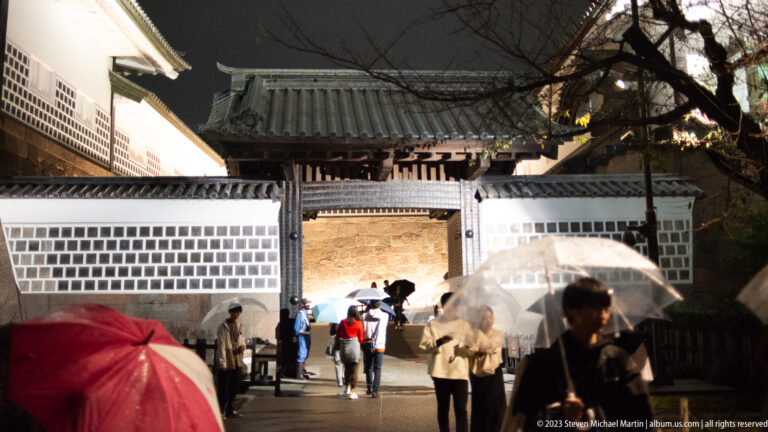 Umbrella-carrying visitors approach a timber castle gate set in white patterned walls at night.