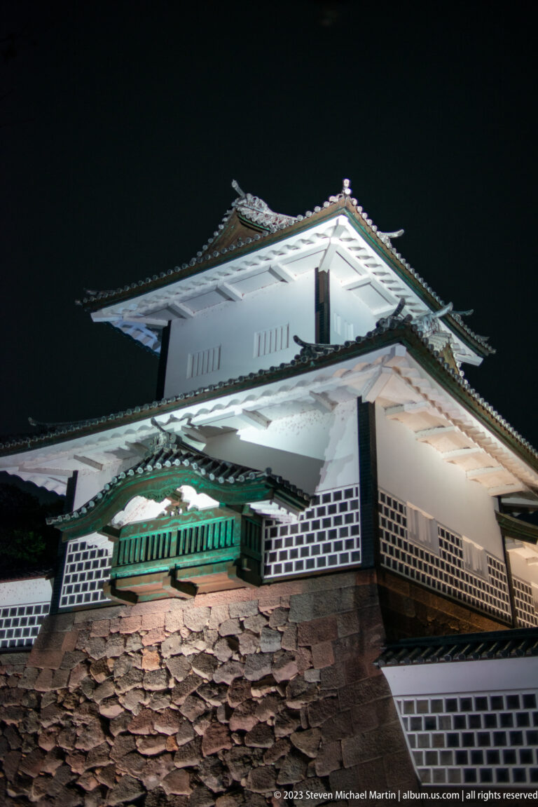 Floodlit white castle turret with layered tiled roofs above a rough stone wall at night.