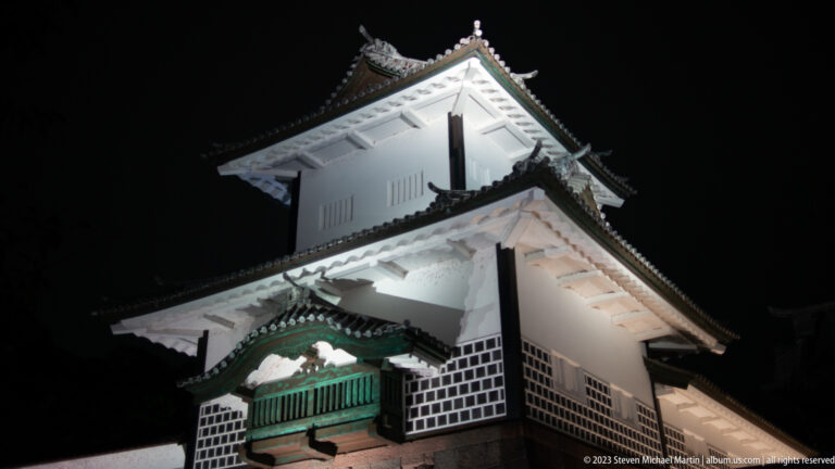 Steep upward view of illuminated white castle walls, dark tiled roofs, and a green railing.
