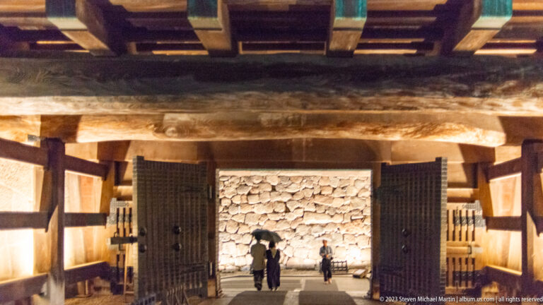Visitors walk through a warmly lit timber castle gate toward a high stone wall.