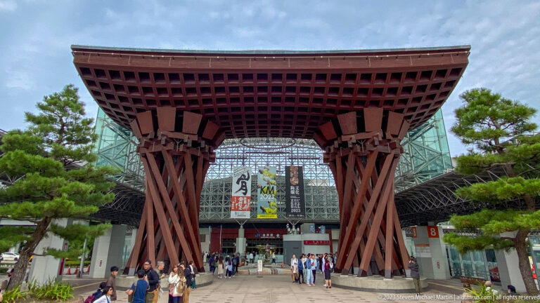 Wider frontal view of Kanazawa Station’s Tsuzumi-mon Gate with pedestrians and trees.
