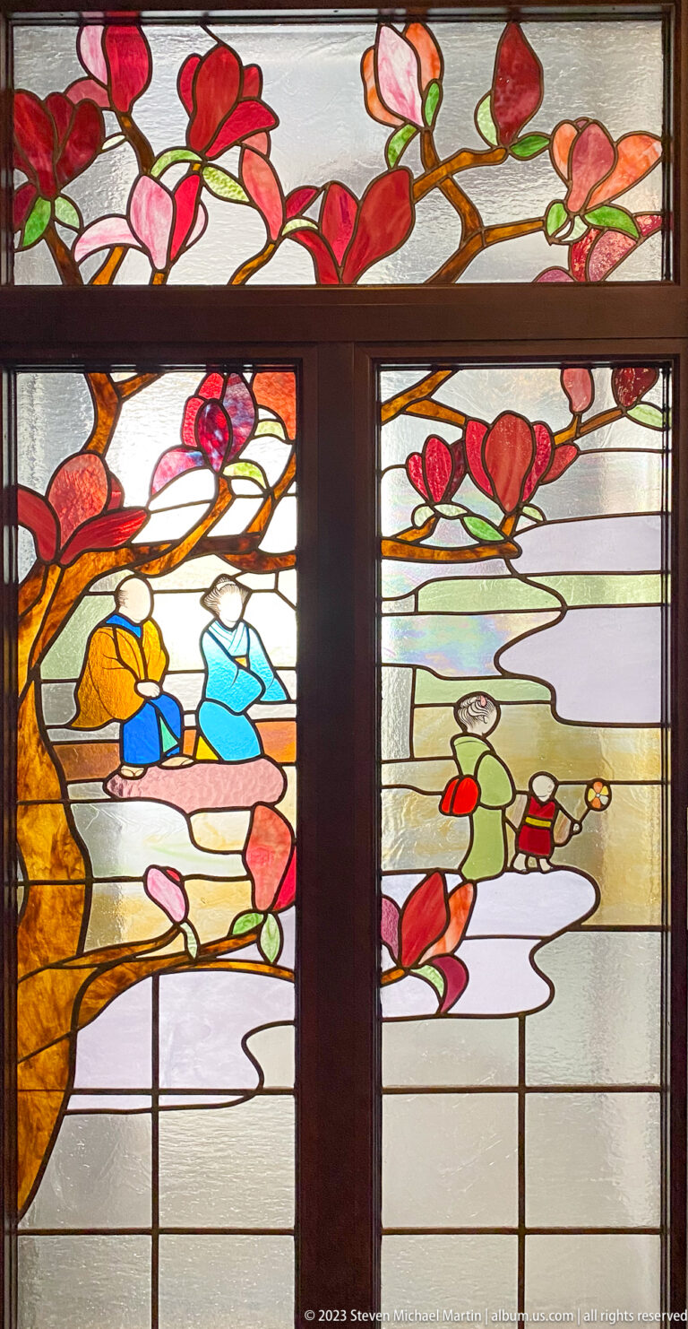 Stained-glass panel with pink magnolia branches and small figures in a stylized landscape.