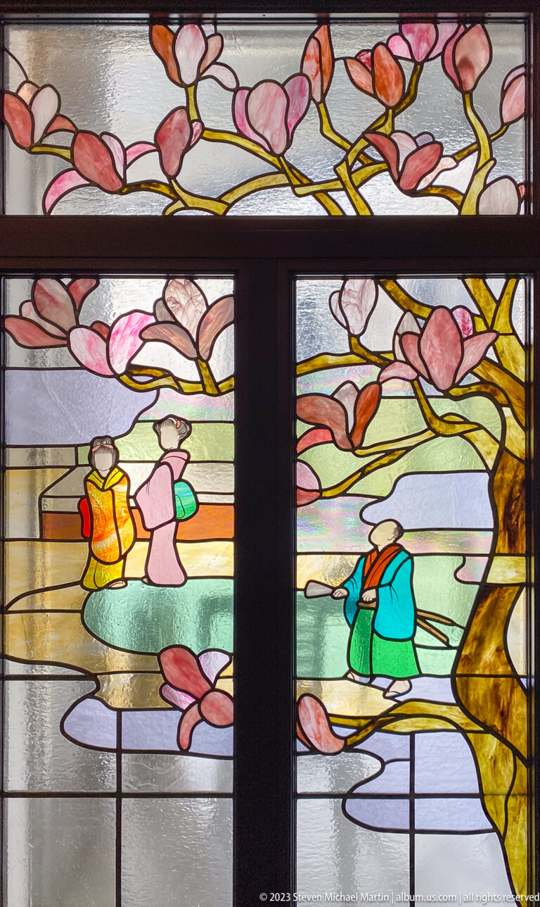 Stained-glass panel showing colorful figures beneath arching pink flowering branches in a carefully framed documentary view.