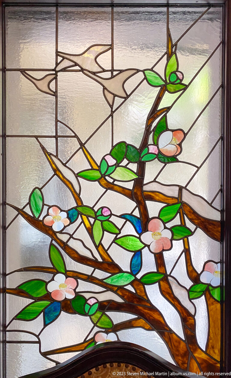 Close-up stained-glass design of brown branches, green leaves, and pale blossoms in a carefully framed documentary view.