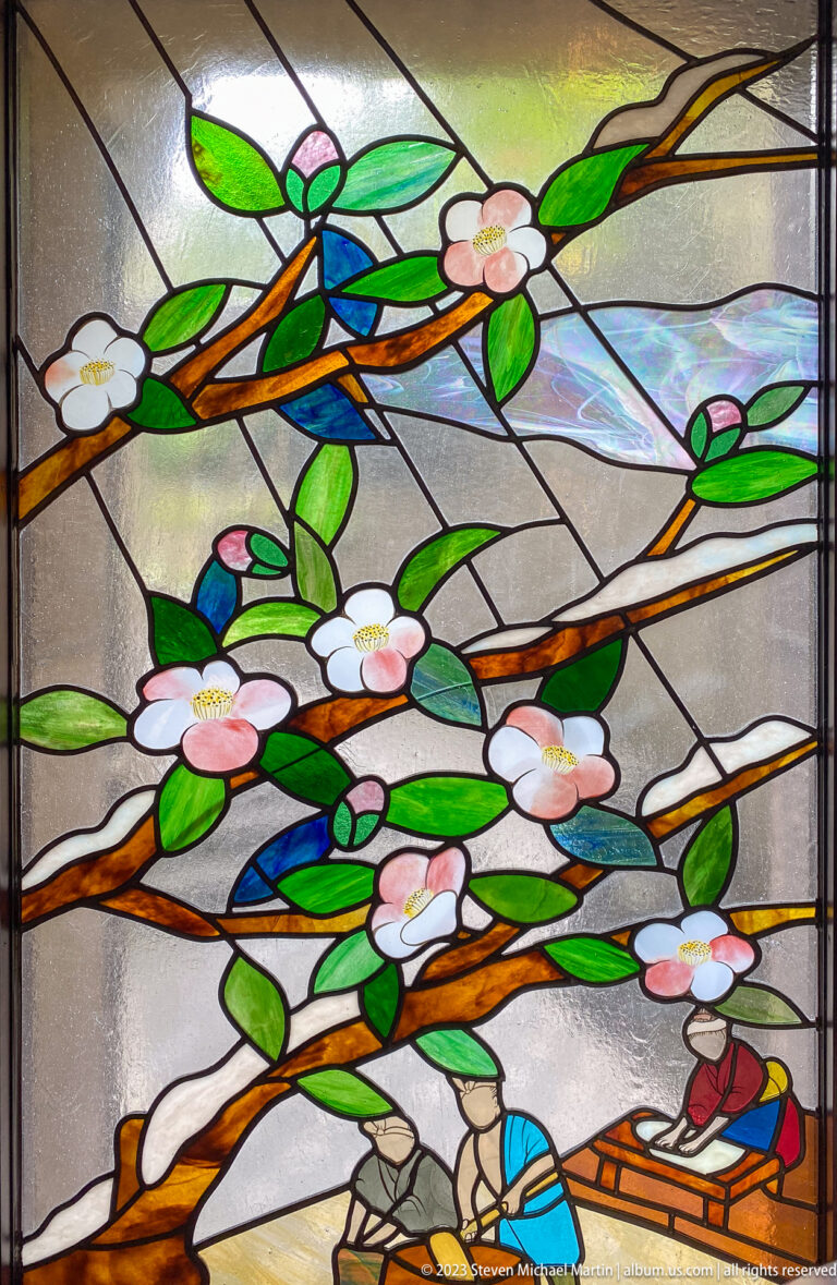 Stained-glass panel with white blossoms, green leaves, and small seated figures below.
