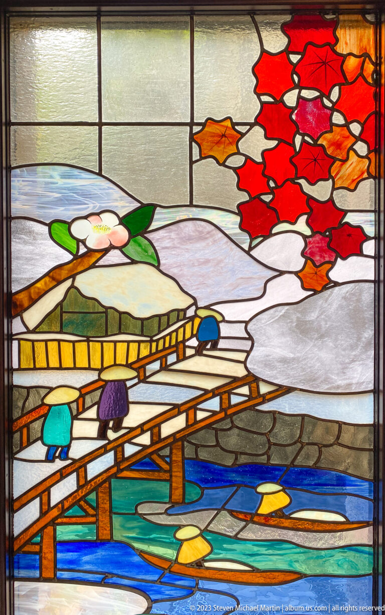 Stained-glass autumn scene with two figures crossing a wooden bridge beneath red maple leaves.