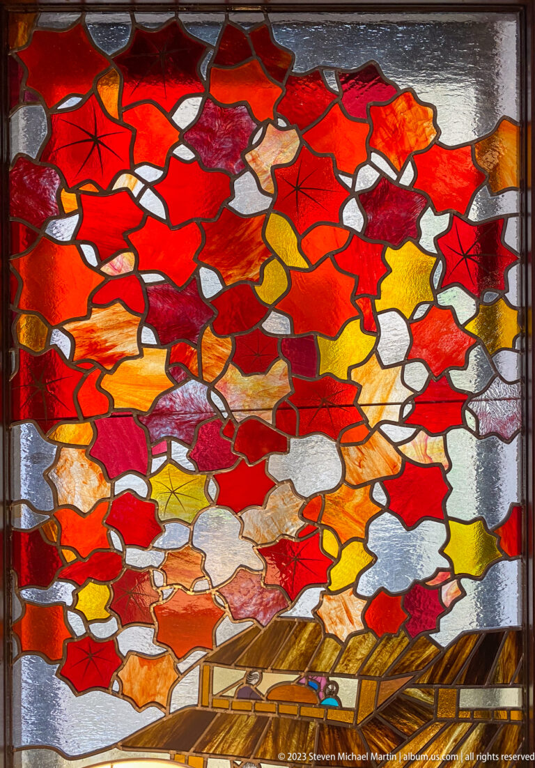 Stained-glass panel dominated by red and orange maple leaves above a small wooden bridge.