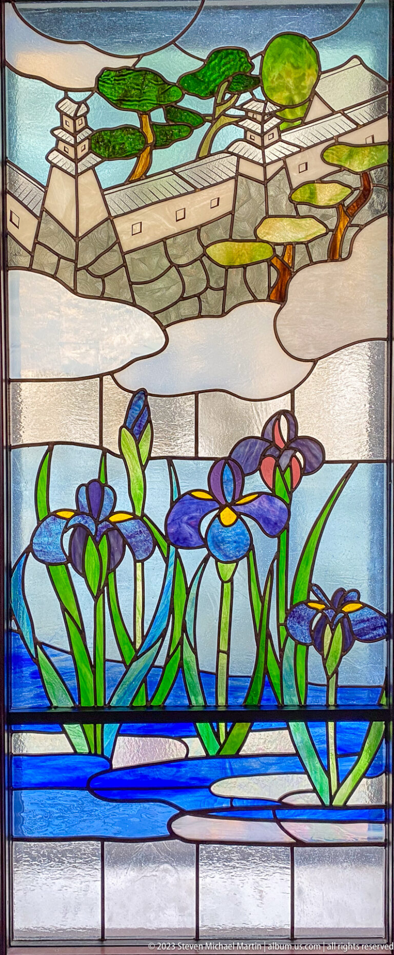 Stained-glass panel with purple irises in blue water before a pale architectural landscape.
