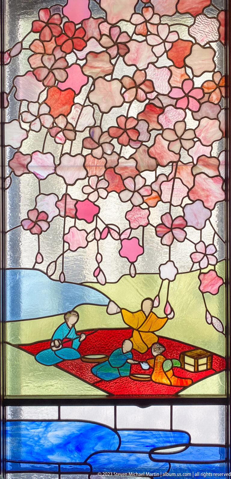 Stained-glass spring scene with pink cherry blossoms above three figures seated in a boat.