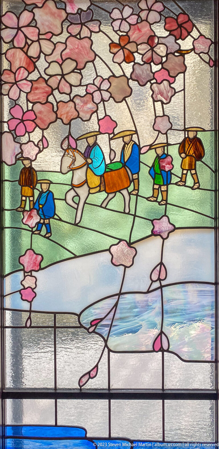 Stained-glass scene of figures walking and riding horses beneath pink cherry blossoms.