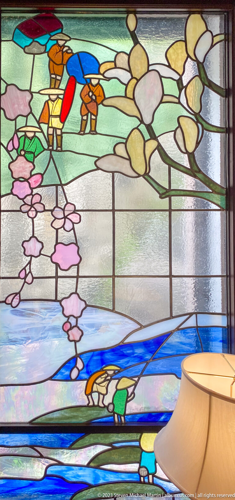Stained-glass panel with wisteria blossoms, a boat, and figures beside blue water.