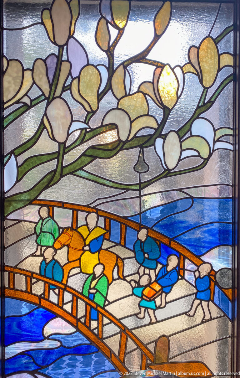 Stained-glass panel showing a crowded wooden bridge beneath pale wisteria blossoms in a carefully framed documentary view.