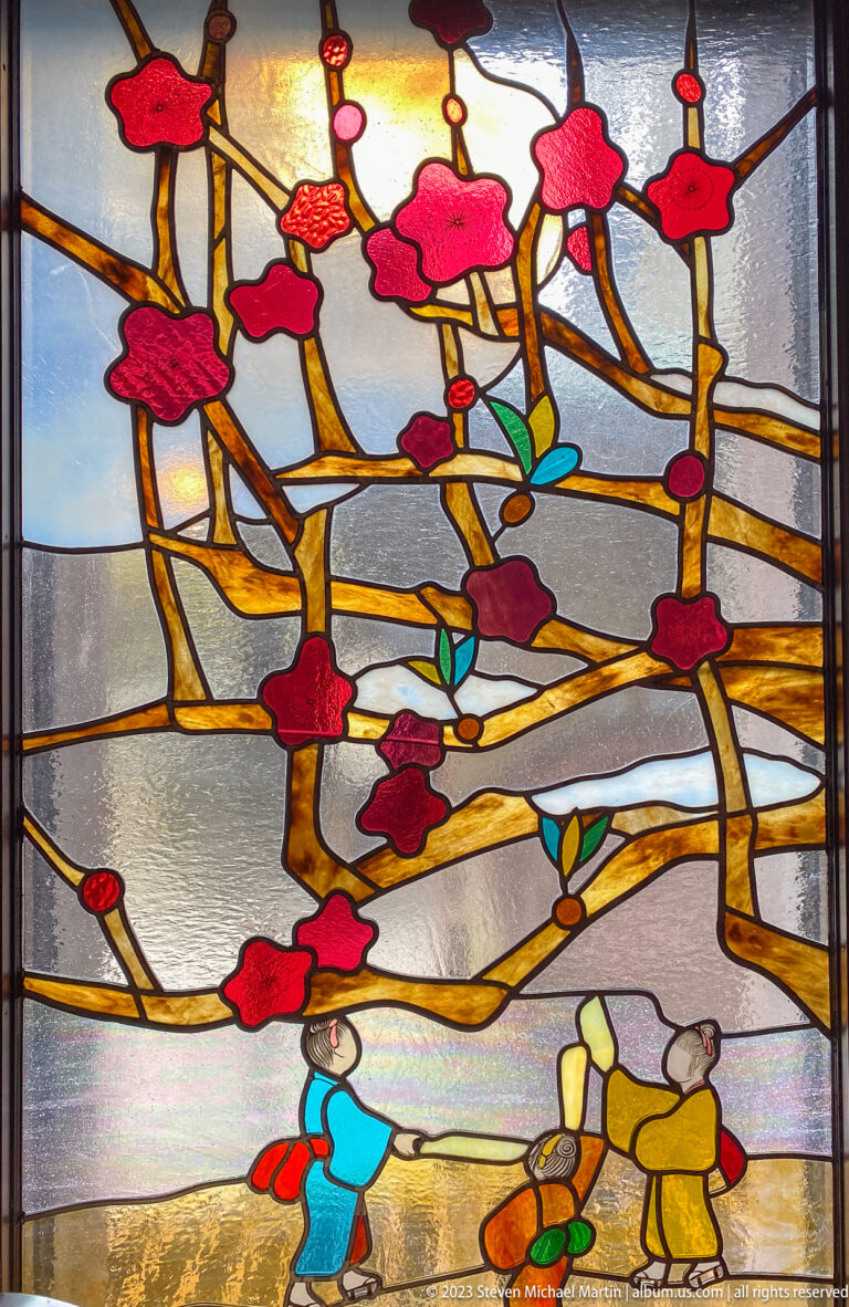 Stained-glass panel of red blossoms above a woman and a stone garden lantern.