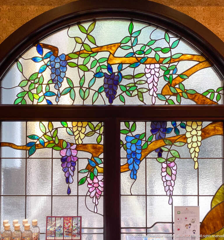Arched stained-glass window with grapevines, green leaves, and multicolored grape clusters in a carefully framed documentary view.