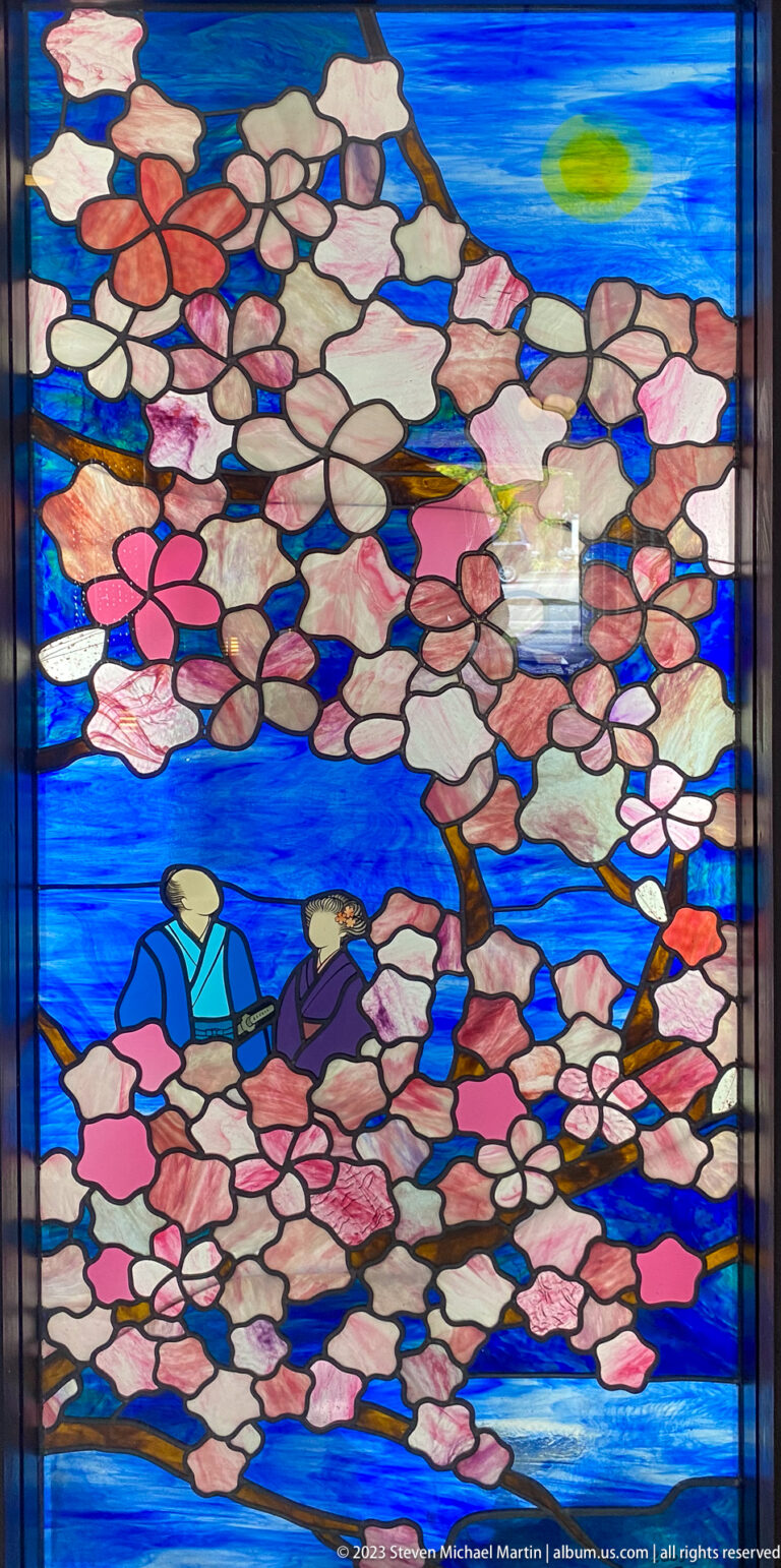 Stained-glass scene with two figures beneath pink blossoms against vivid blue water or sky.