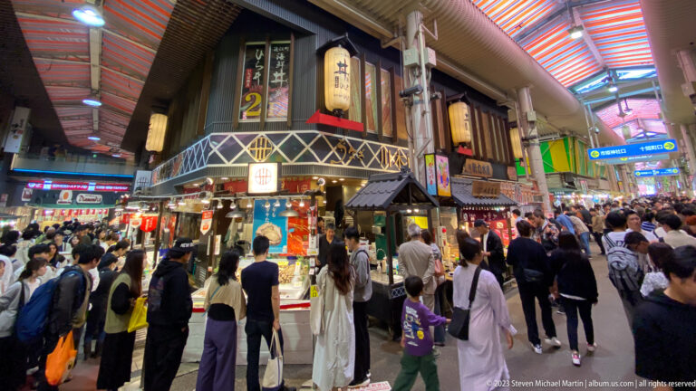 Crowded covered market arcade with shoppers, food stalls, hanging signs, and colorful roof panels.