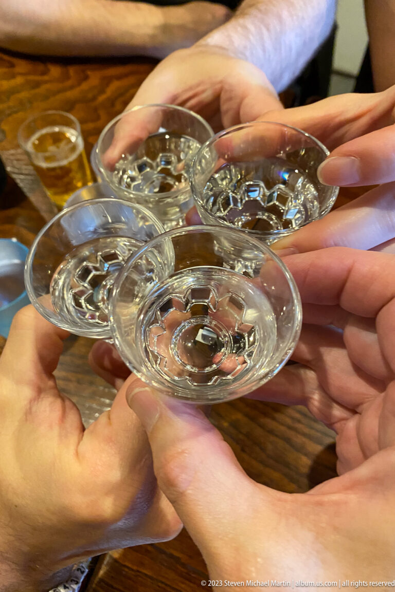 Several hands clink shallow clear glass sake cups together over a restaurant table.