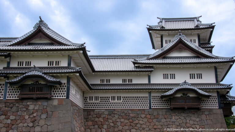 White multi-level Kanazawa Castle buildings with gabled roofs and projecting windows above colorful stonework.