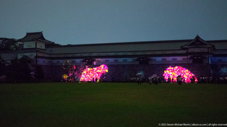 Kanazawa Castle walls and turrets behind two glowing multicolored installations on a dark lawn at dusk.