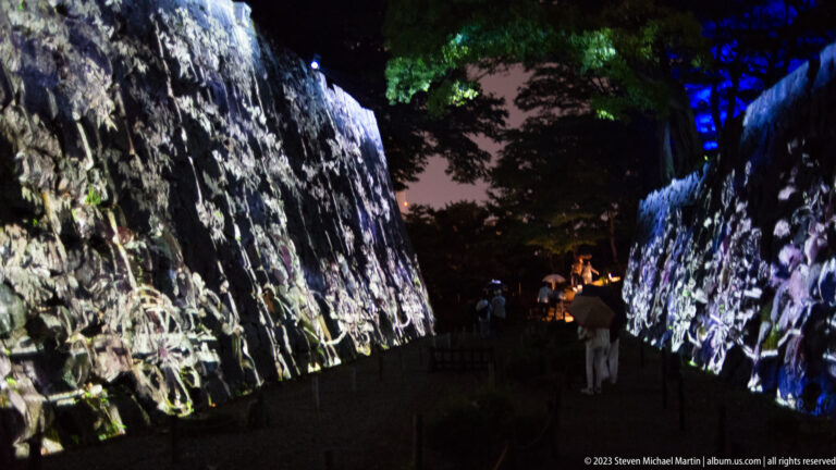 Visitors walk between projection-covered stone walls beneath blue and green lighting.