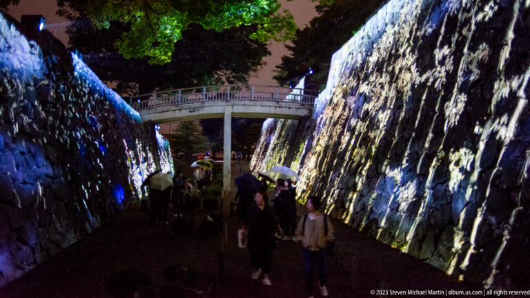 Visitors walk through a projection-lit stone corridor beneath a bridge.