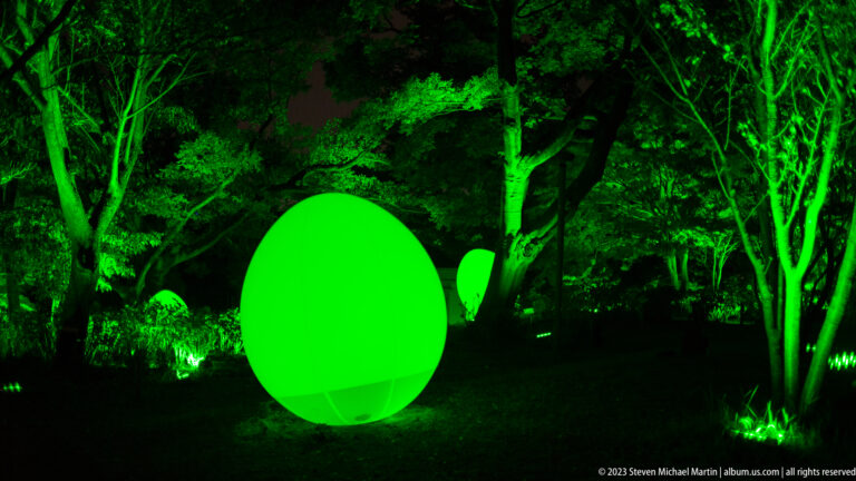 Large glowing green responsive ovoid sculpture among trees, with a smaller matching form behind.