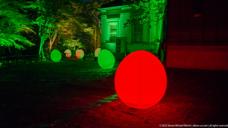 Large red glowing responsive ovoid sculpture near a green-lit historic building and smaller ovoids.