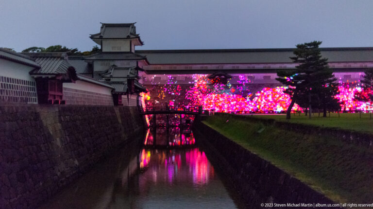 Pink and red illuminated trees reflect in a narrow moat beside Kanazawa Castle walls and tiled roofs.