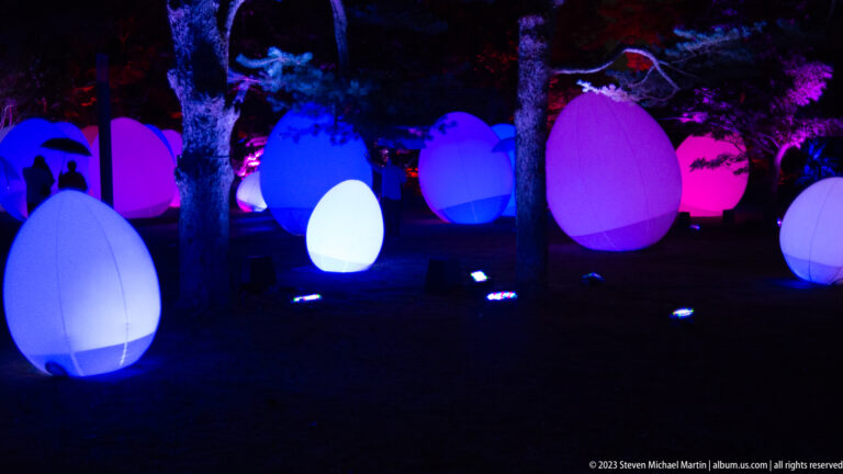Cluster of multicolored glowing responsive ovoid installations scattered among trees.