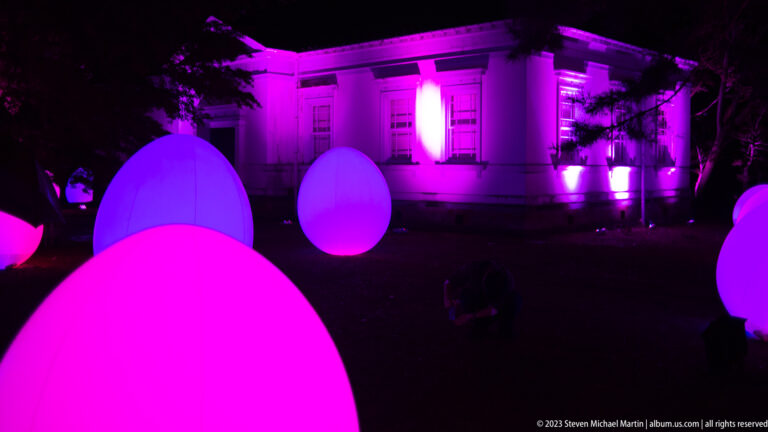 Pink and violet responsive glowing ovoids in front of a purple-lit historic building.