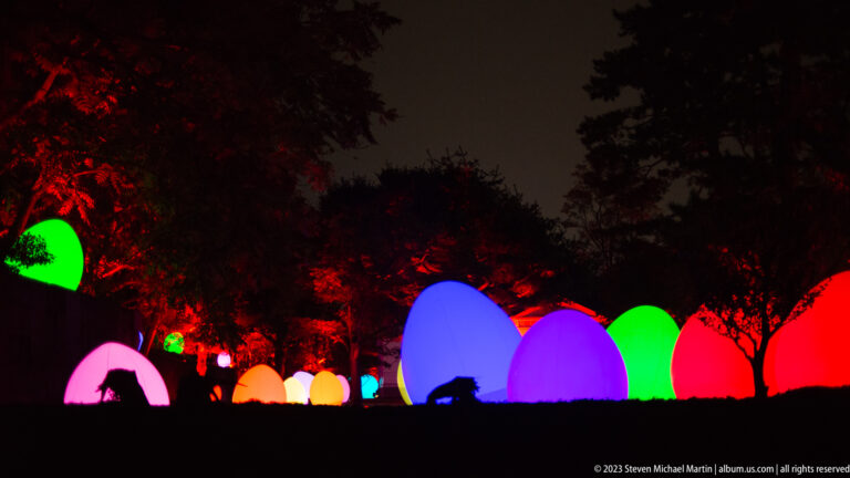Low-angle row of multicolored responsive glowing ovoids beneath red-lit trees.