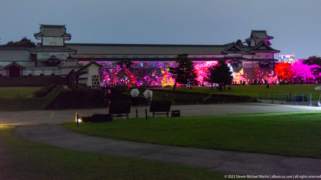 Curving path and lawn lead toward brightly illuminated plantings below Kanazawa Castle walls and towers.