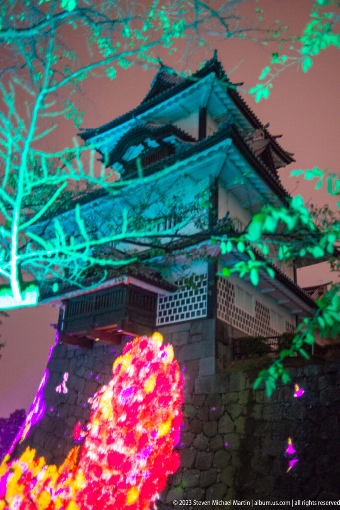 Castle turret and walls behind a bright pink-red installation and cyan-lit branches.