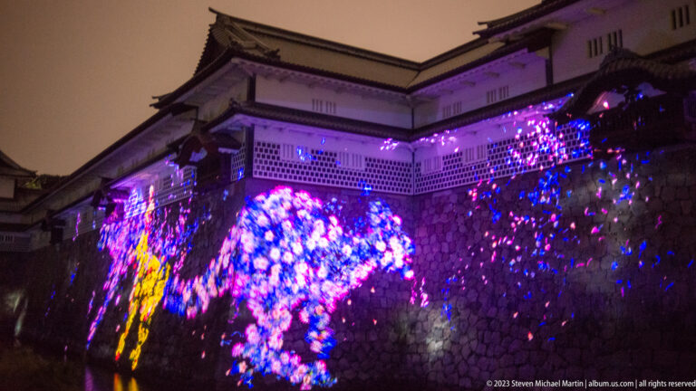 Multicolored blossom-like projections cover a stone wall below Kanazawa Castle buildings.