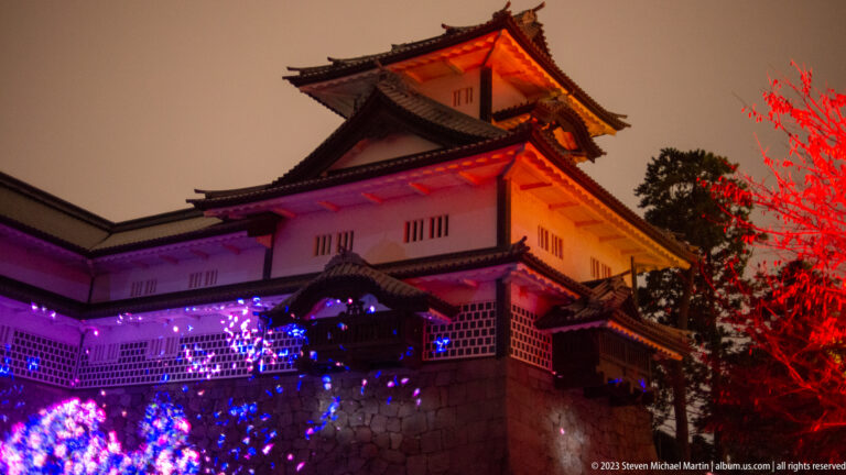 Kanazawa Castle turret lit orange and red above violet-blue projections on the stone wall.