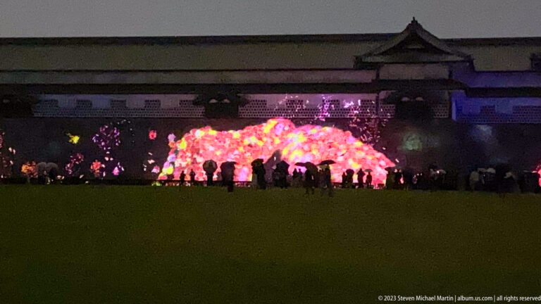 Visitors stand before a glowing pink installation below Kanazawa Castle walls at night.